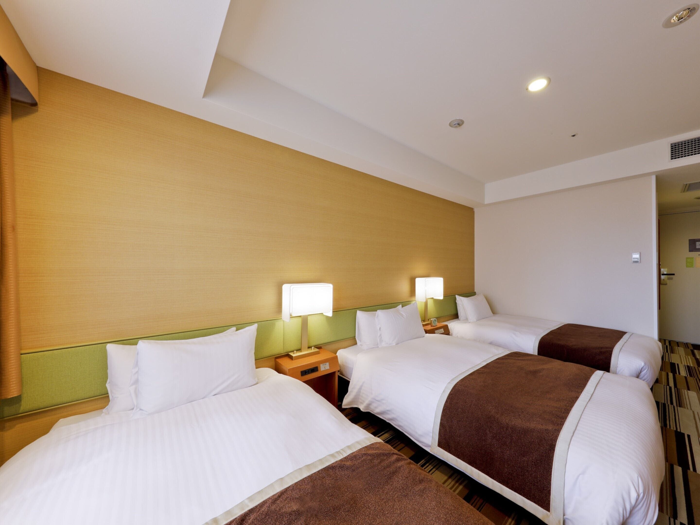 A hotel room with three beds, each with white bedding and brown throw blankets. There are bedside tables with lamps and a visible door to another room.