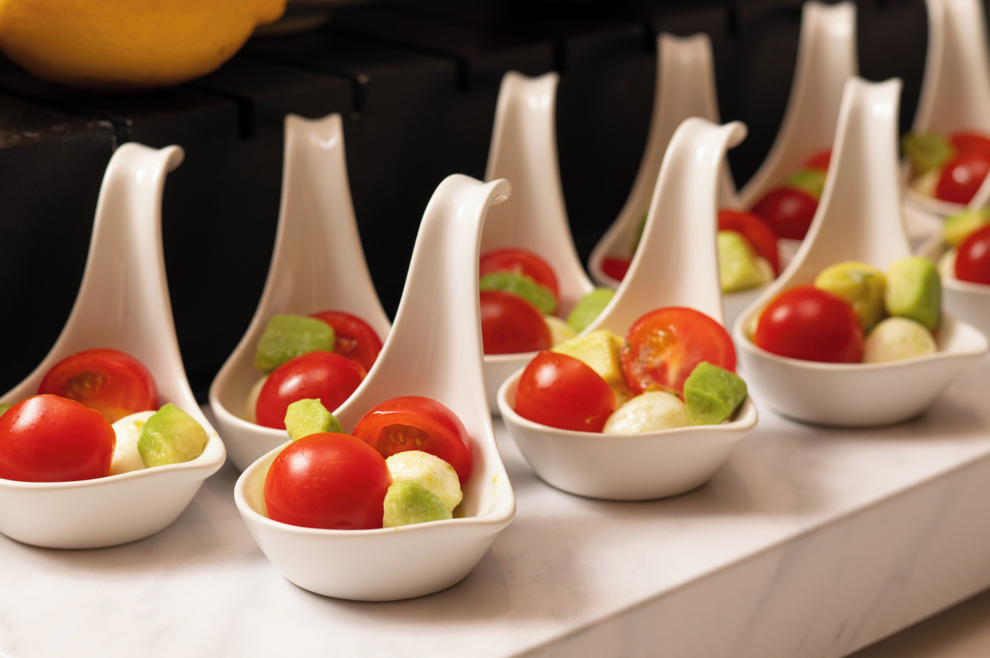 A series of white, spoon-shaped bowls containing cherry tomatoes and avocado slices.