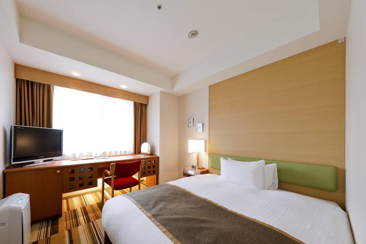A hotel room with a bed, a desk with a television, a chair, a lamp, and a window with curtains.
