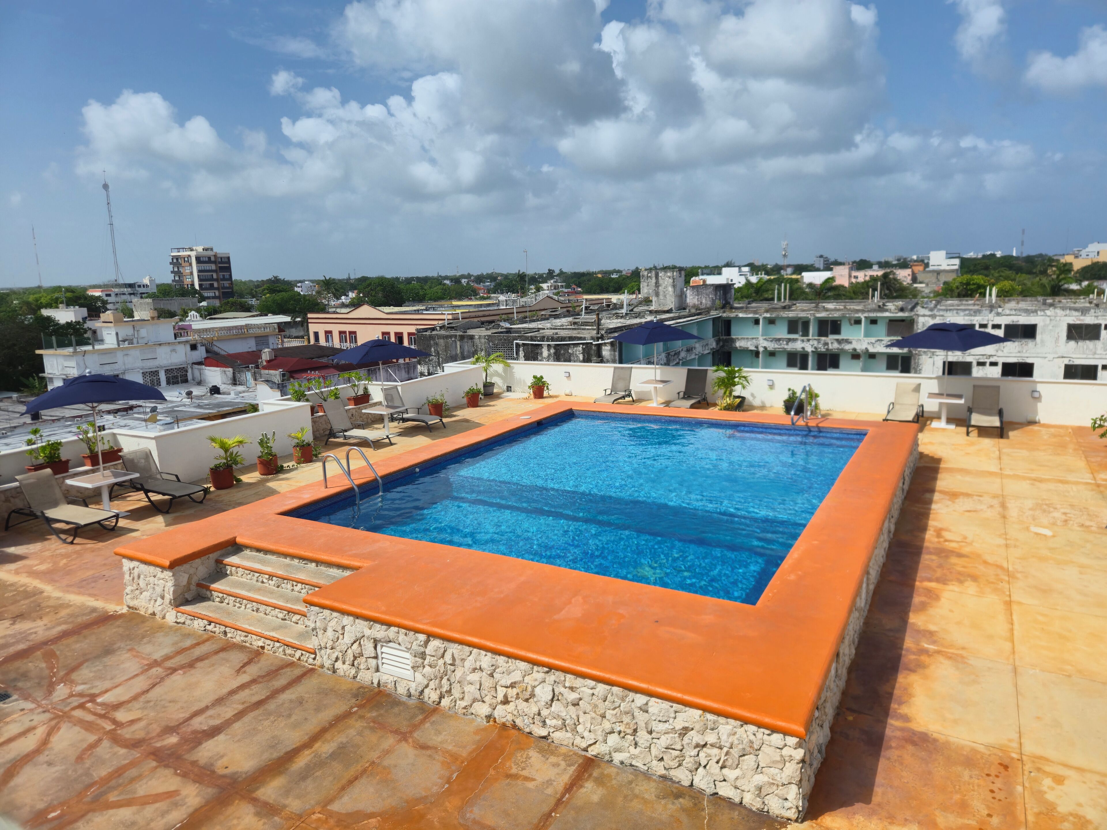 A rooftop swimming pool with a stone border, lounge chairs, and potted plants.