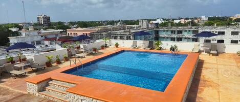 A rooftop swimming pool with a stone border, lounge chairs, and potted plants.