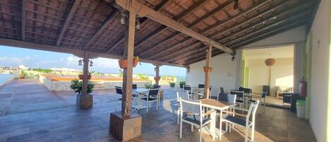 An outdoor patio with stone flooring, wooden tables and chairs, hanging lanterns, and potted plants.
