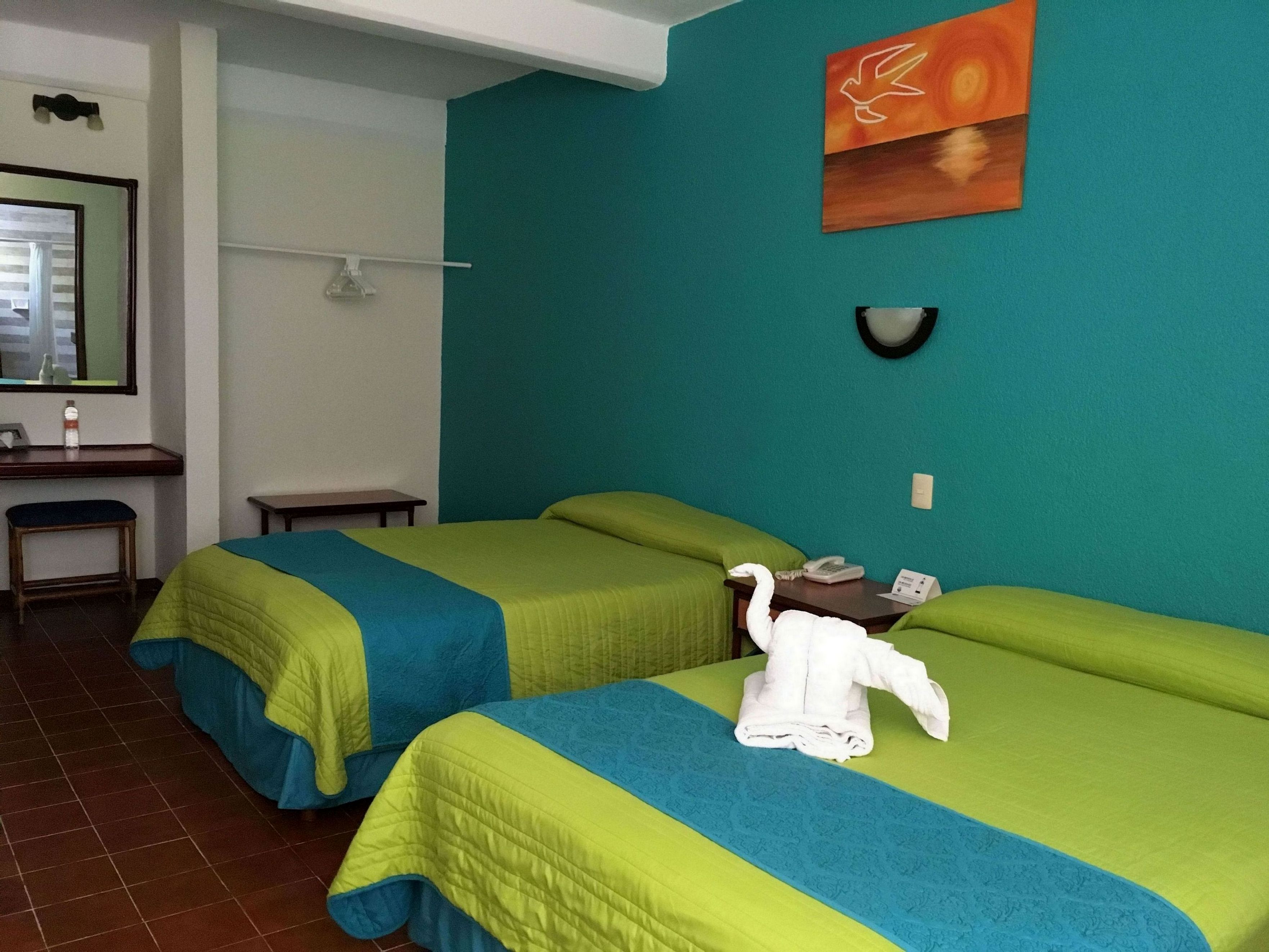 A hotel room with two beds, each with a green and blue bedspread, a small table with a telephone, and a wall-mounted light fixture.
