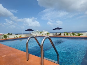 A swimming pool with metal ladders, two umbrellas, and a cruise ship in the background. - Hotel Plaza Cozumel (Cozumel)