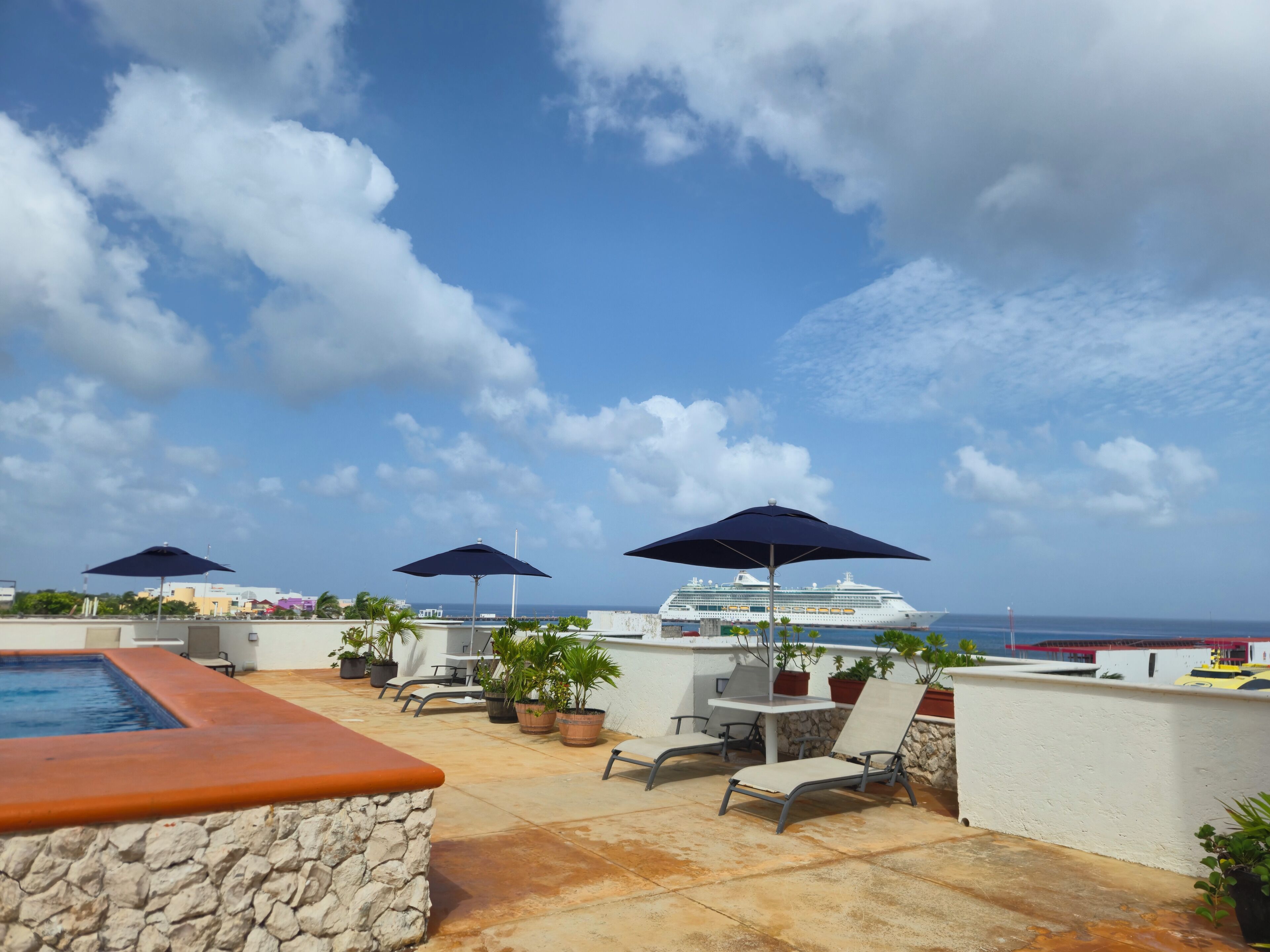 A rooftop terrace with a pool, lounge chairs, umbrellas, and a cruise ship in the background.