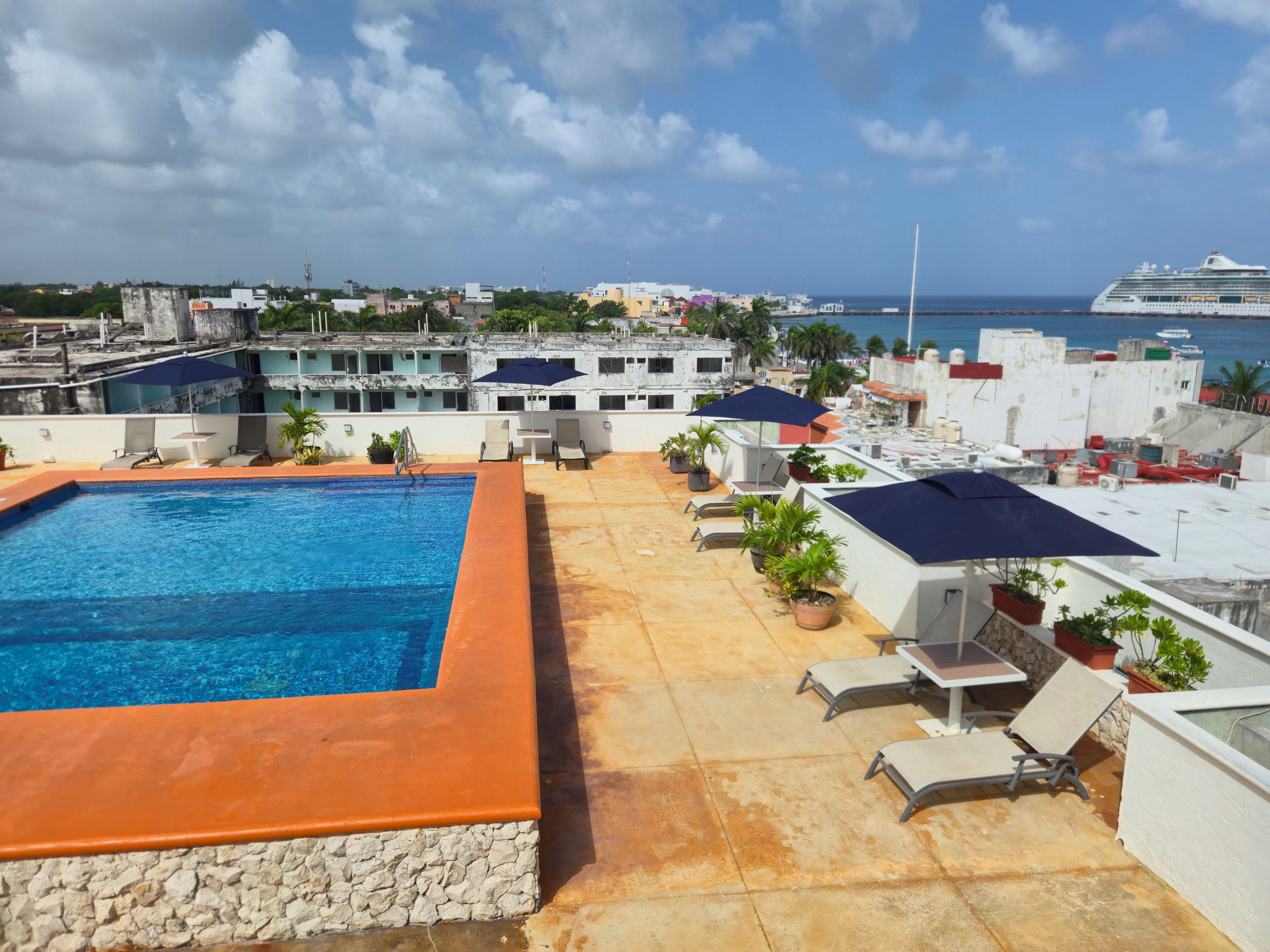 Une piscine sur le toit, dotée de chaises longues et de parasols, avec une vue sur la mer et des bâtiments en arrière-plan.