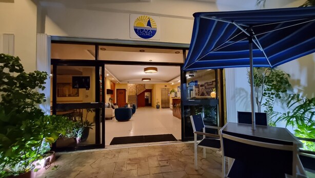 A hotel entrance with a sign reading "Plaza Cozumel," a covered seating area with a table and chairs, and potted plants around. - Hotel Plaza Cozumel (Cozumel)