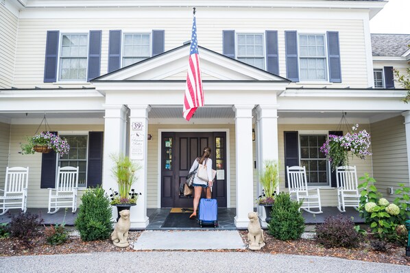 A two-story building with a porch, American flag, and two white rocking chairs.