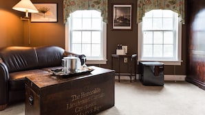 A room with a brown leather sofa, a wooden chest labeled "The Honorable Lieutenant Captain Greenwood's Bay," and two windows with floral curtains. - The Reluctant Panther Inn and Restaurant (Manchester)