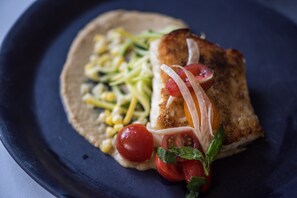 Um prato com peixe grelhado, milho, abobrinha, tomates cereja e manjericão.