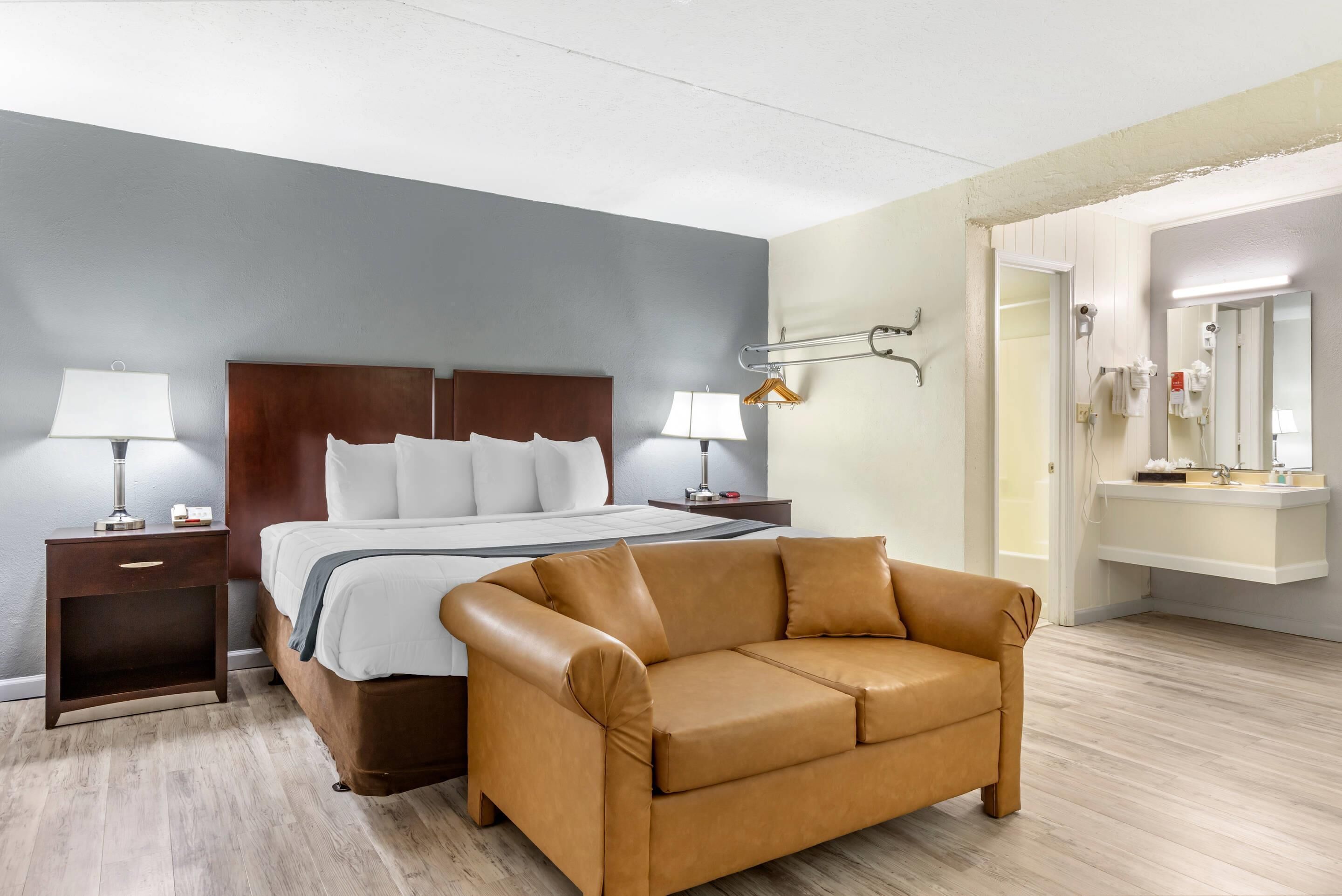 A hotel room with a bed, a brown leather sofa, a nightstand with a lamp, a wall-mounted towel rack, and a bathroom with a sink and mirror.