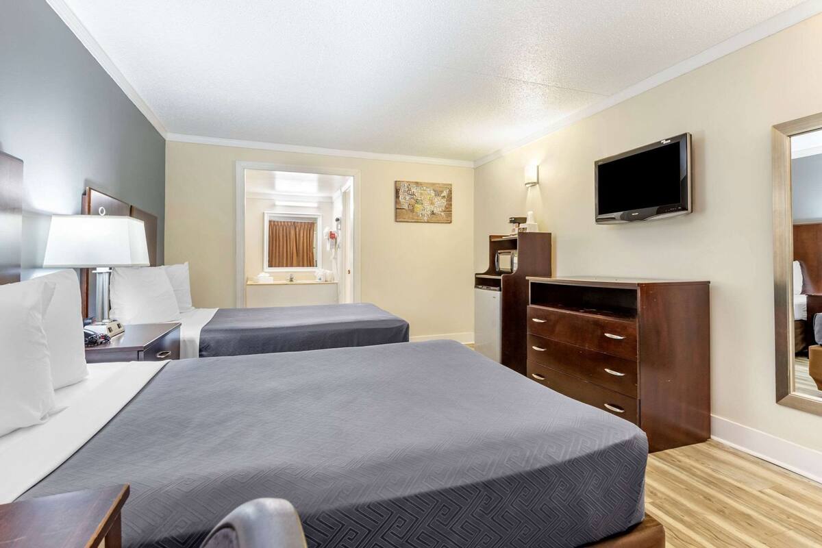 Habitación de hotel con dos camas, televisión, escritorio con lámpara, cómoda y baño visible a través de una puerta abierta.