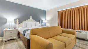 A hotel room with a bed, a nightstand with a lamp, a sofa, and a window with curtains. - Econo Lodge Conway Central (Conway)