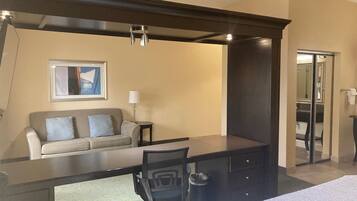 A hotel room with a bed, a desk, a chair, a sofa, a lamp, a trash can, and a framed picture on the wall.