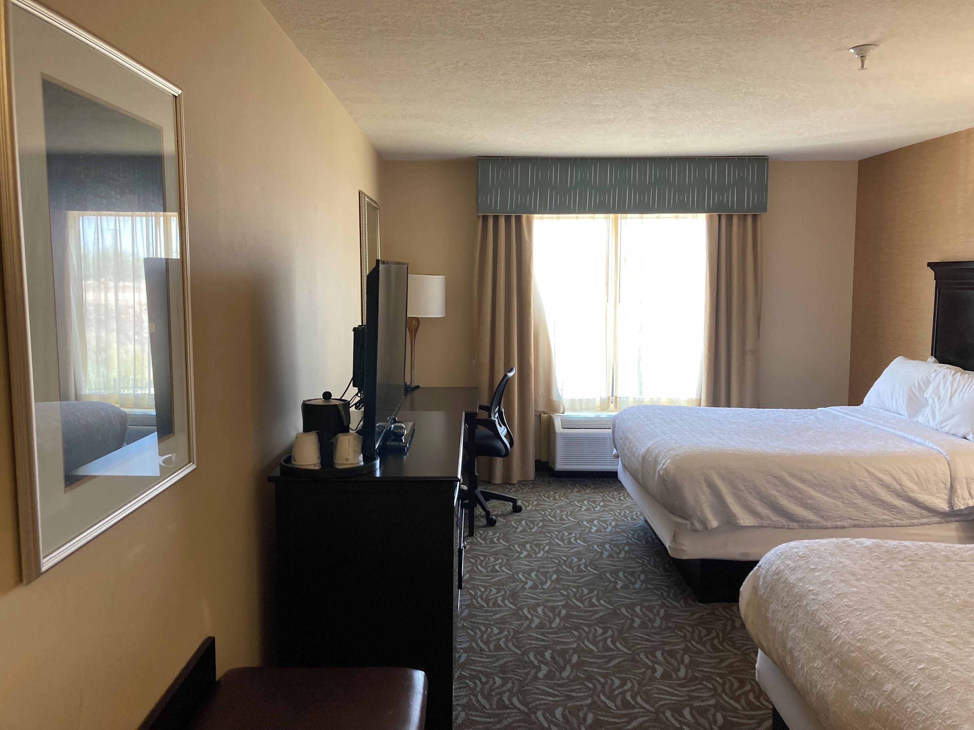 A hotel room with two beds, a desk with a lamp, a chair, and a large mirror.