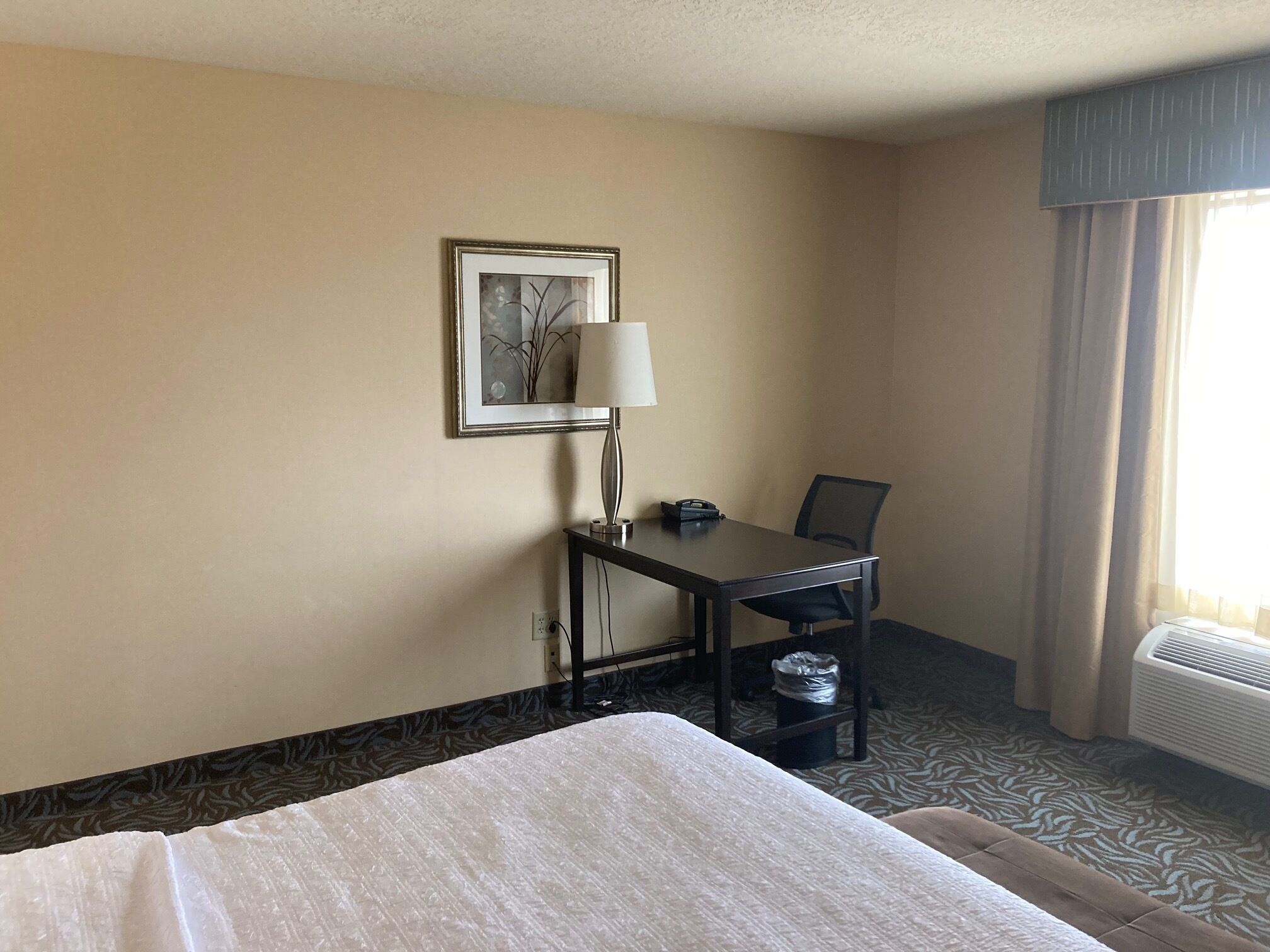 A hotel room with a bed, a desk with a lamp, a chair, a picture on the wall, and a window with curtains.