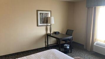 A hotel room with a bed, a desk with a lamp, a chair, a picture on the wall, and a window with curtains.
