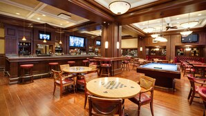 A bar area with wooden flooring, multiple bar stools, a pool table, and a television mounted on the wall.