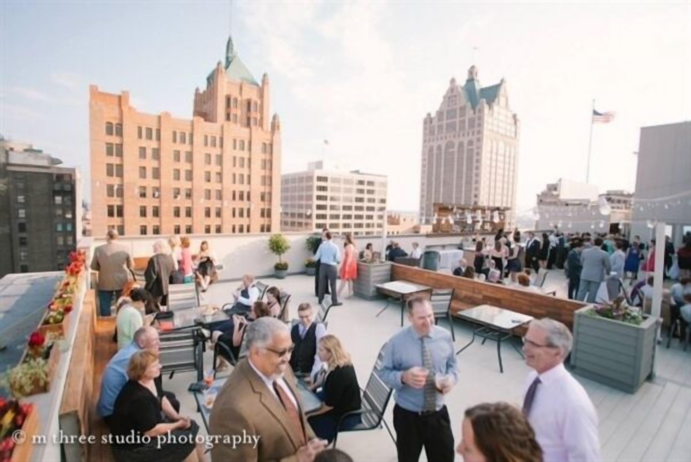 A rooftop event with people seated and standing, surrounded by modern buildings and a clear sky.
