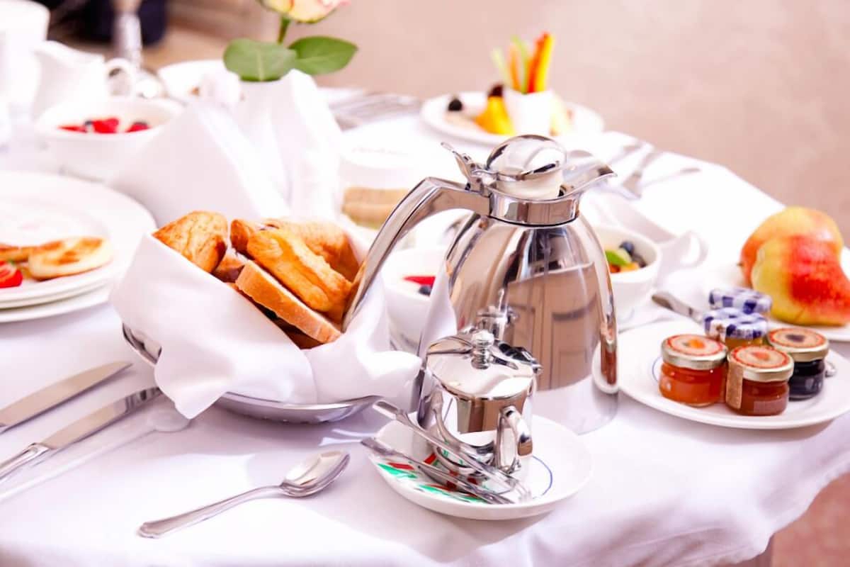 Ein Frühstücksarrangement mit einer silbernen Kaffeekanne, Croissants und verschiedenen Marmeladen auf einer weißen Tischdecke.