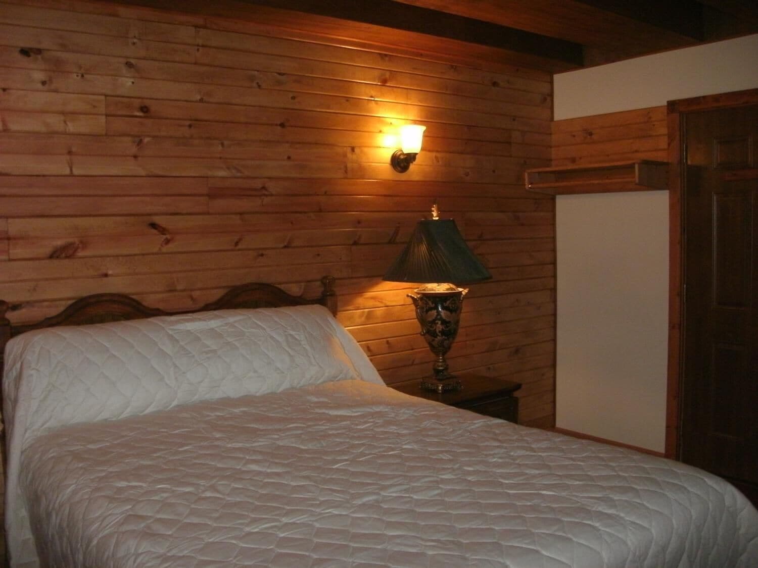 Una stanza con rivestimento in legno, un letto, un comodino con una lampada e una porta.