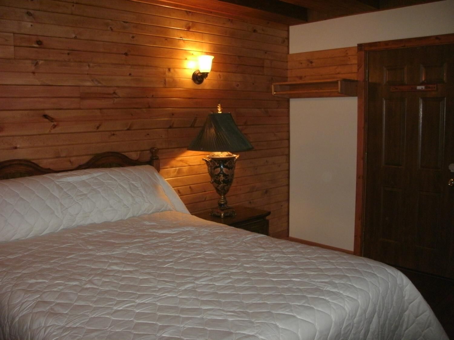 Een slaapkamer met een houten wand, een bed met een gewatteerde spreideken, een bedlamp en een houten deur.