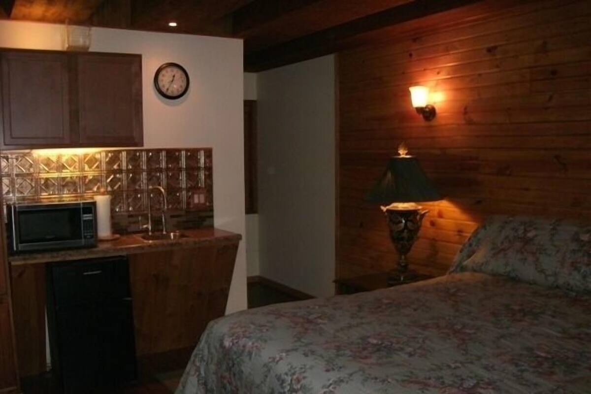 A compact room with a bed, a small kitchenette, a clock, and a wall-mounted lamp.
