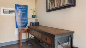 A hotel room with a wooden desk, a potted plant, a framed artwork, and a promotional poster.