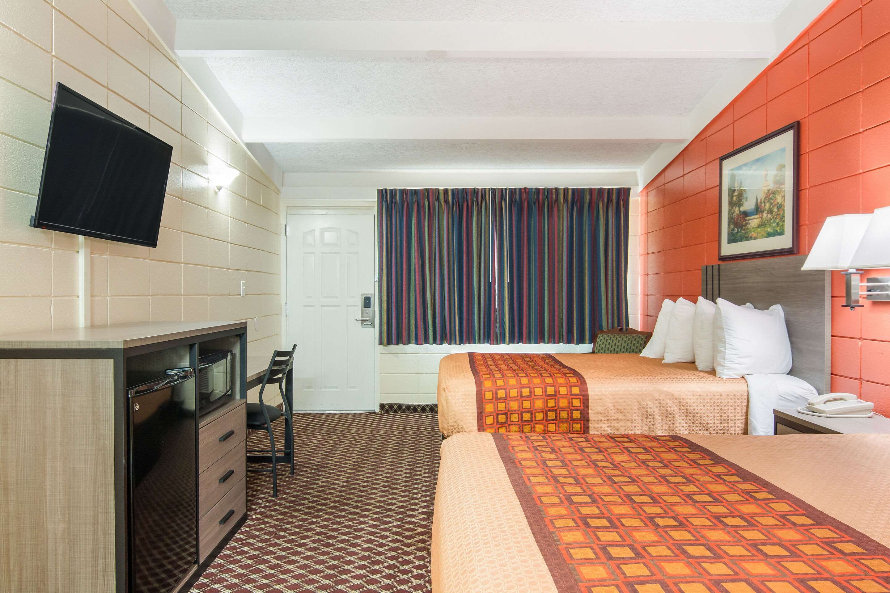 A hotel room with a bed, a desk with a TV, a chair, a window with curtains, and a painting on the wall.