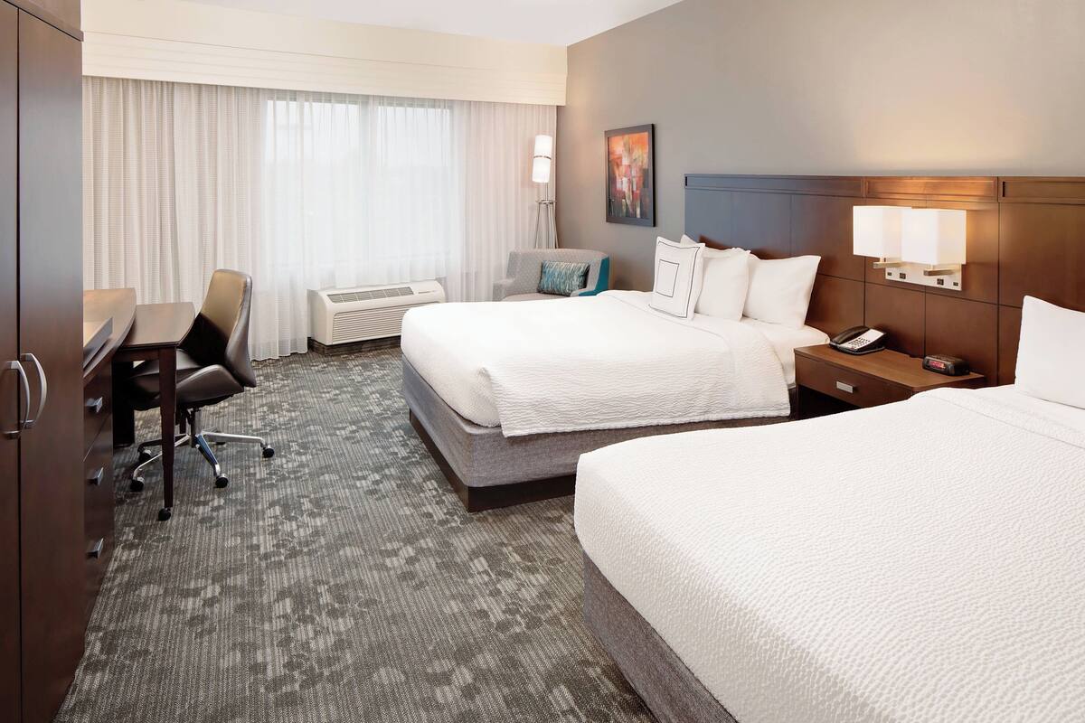 A hotel room with two beds, a desk, a chair, a lamp, and a painting on the wall.