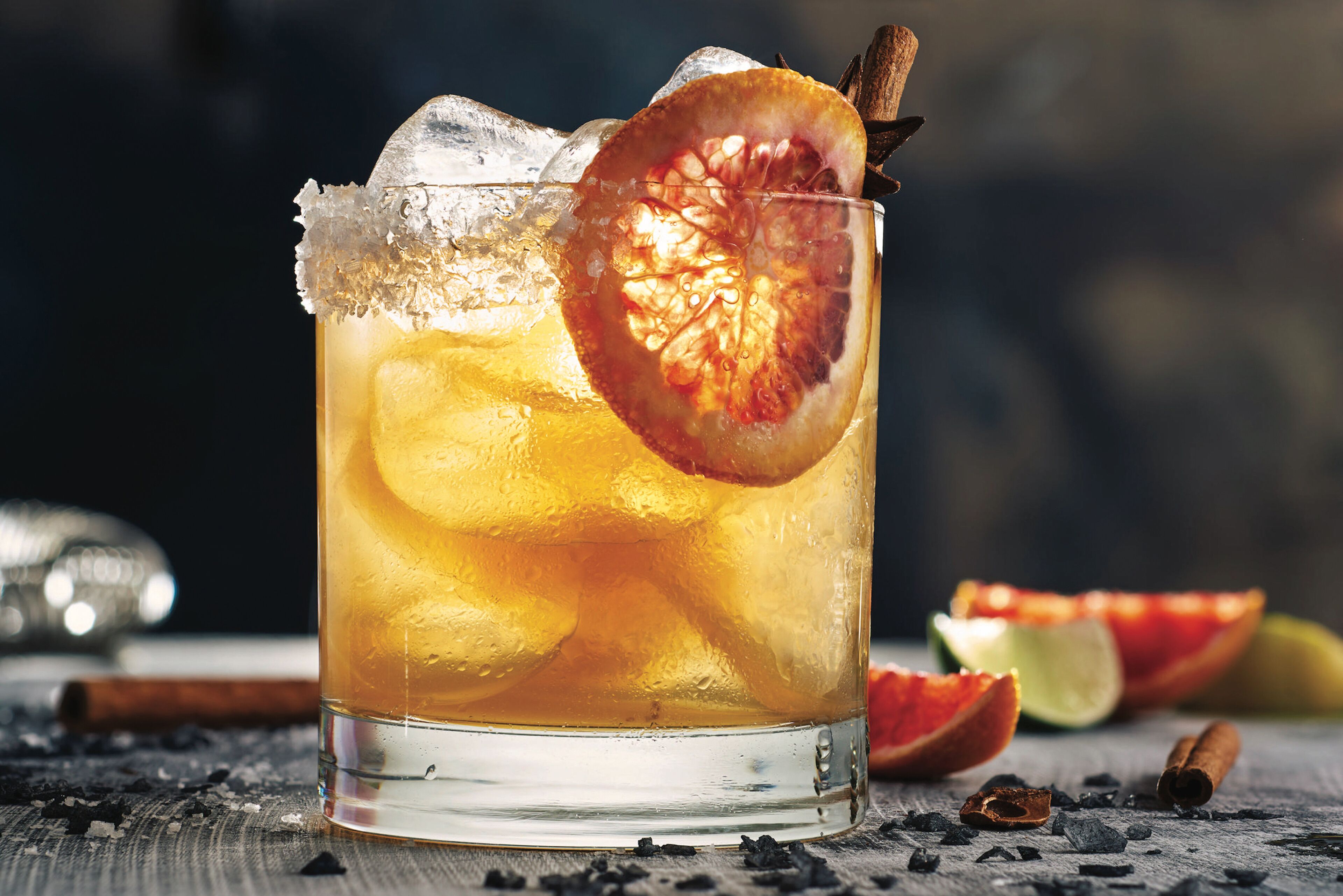 A glass of iced beverage with a slice of blood orange and a cinnamon stick, surrounded by scattered spices and a half-cut fruit on a wooden surface.