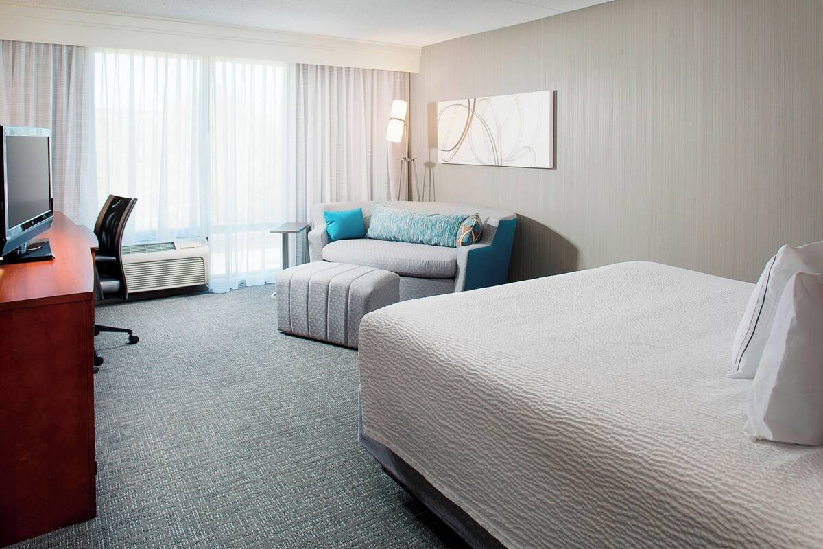 A hotel room with a large bed, a smaller sofa, a desk with a computer, and a window with curtains.