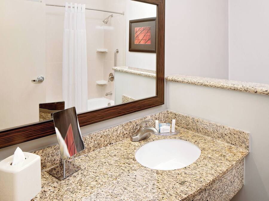 A bathroom with a granite countertop, a white sink, a mirror, a framed picture, and a towel dispenser.
