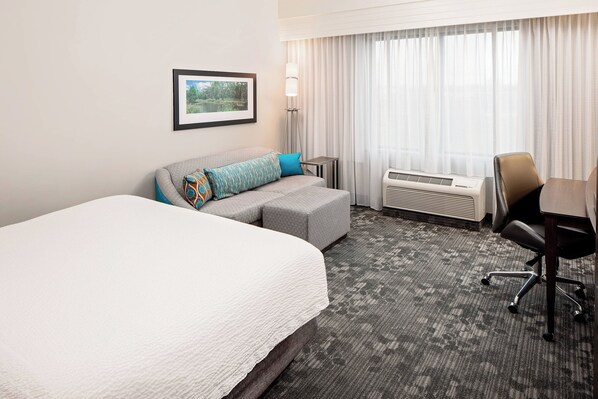 A hotel room with a large bed, a gray sofa with colorful pillows, a desk with a chair, a standing lamp, and a window with curtains.