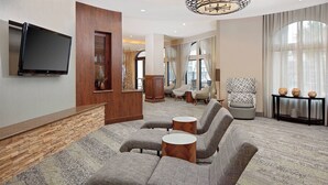 A modern hotel lobby with a stone accent wall, a flat-screen TV, a seating area with grey upholstered sofas and round wooden side tables, and a large window with sheer curtains.