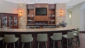 A bar area with a granite countertop, bar stools, a television mounted on a stone wall, and shelves with bottles and glasses.