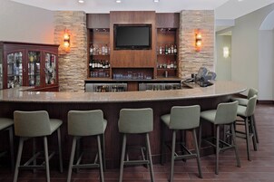 A bar area with a granite countertop, bar stools, a television mounted on a stone wall, and shelves with bottles and glasses.