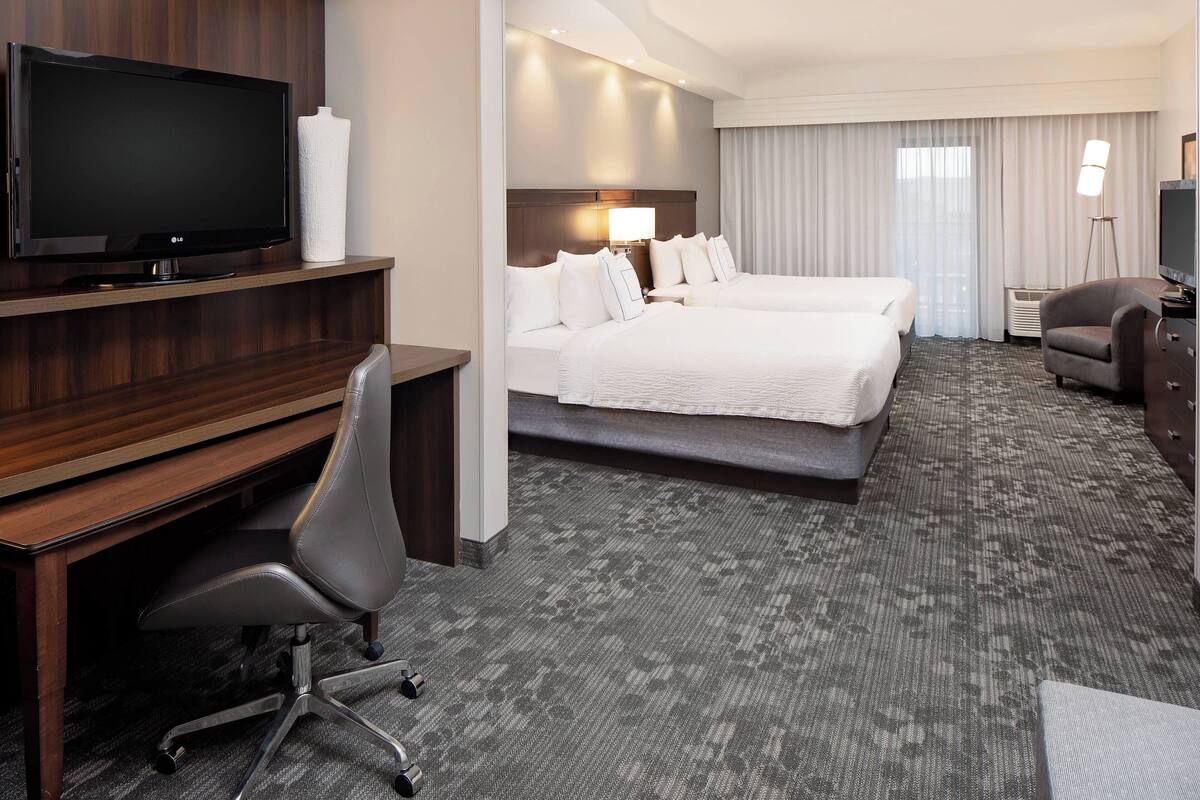 A hotel room with a flat-screen TV, a desk with a chair, two beds, a lamp, and a TV on a stand.