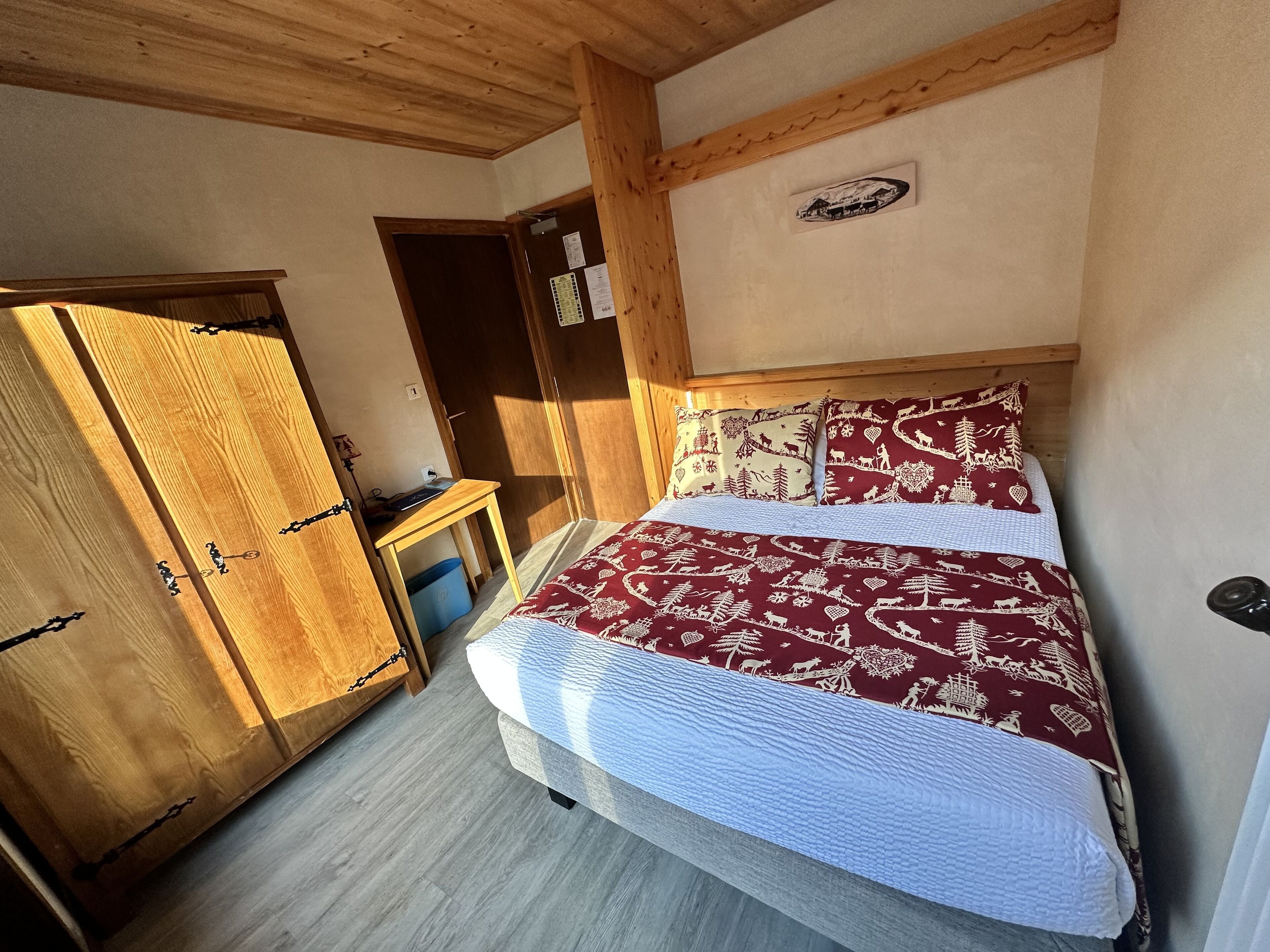 A bedroom with a bed, wooden wardrobe, and a small table with a chair.