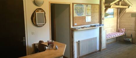 A room with a wooden floor, a dining table with chairs, a radiator, a wooden bench with a cushion, and a wall with a mirror and a bulletin board.