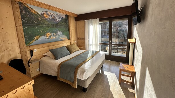 A cozy bedroom with a large bed, a wooden desk, a painting of a mountain landscape, and a balcony with a view of trees. - Le Soly Hotel (Morzine)