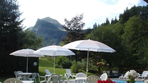 An outdoor seating area with white plastic chairs and tables, white umbrellas, and a pool in the background. - Le Soly Hotel (Morzine)