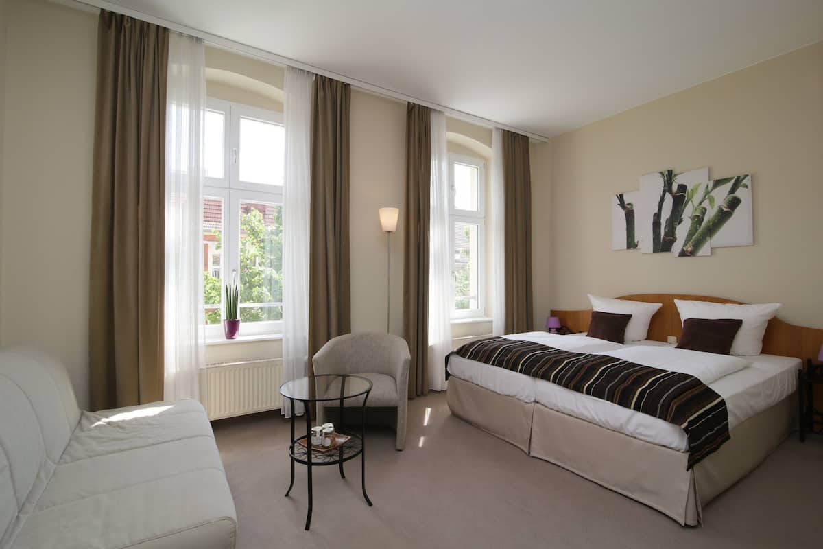 A hotel room with a bed, a chair, a small table, and a window with curtains.