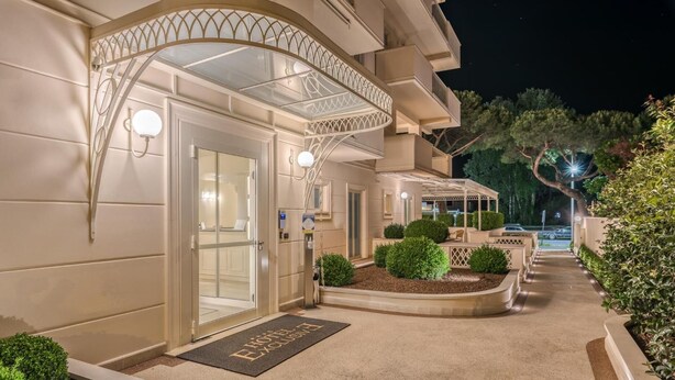 Entrada de hotel con puerta de cristal, zona de estar exterior cubierta y un camino bien iluminado.