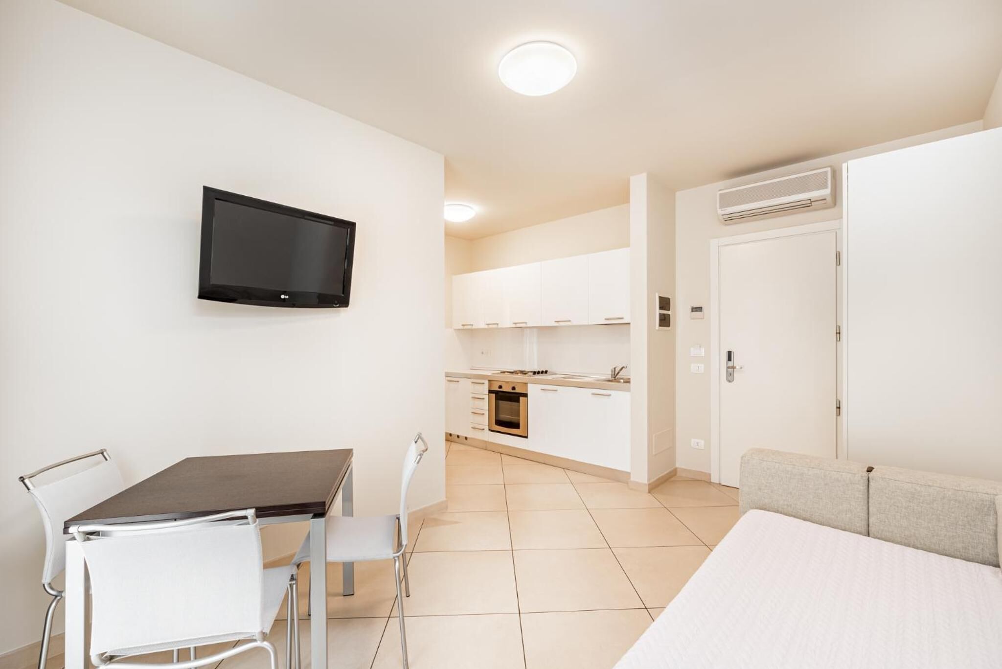 Een compacte kamer met een kitchenette, een eethoek met tafel en stoelen, een flatscreen televisie aan de wand en een bed met een grijze hoofdeinde.