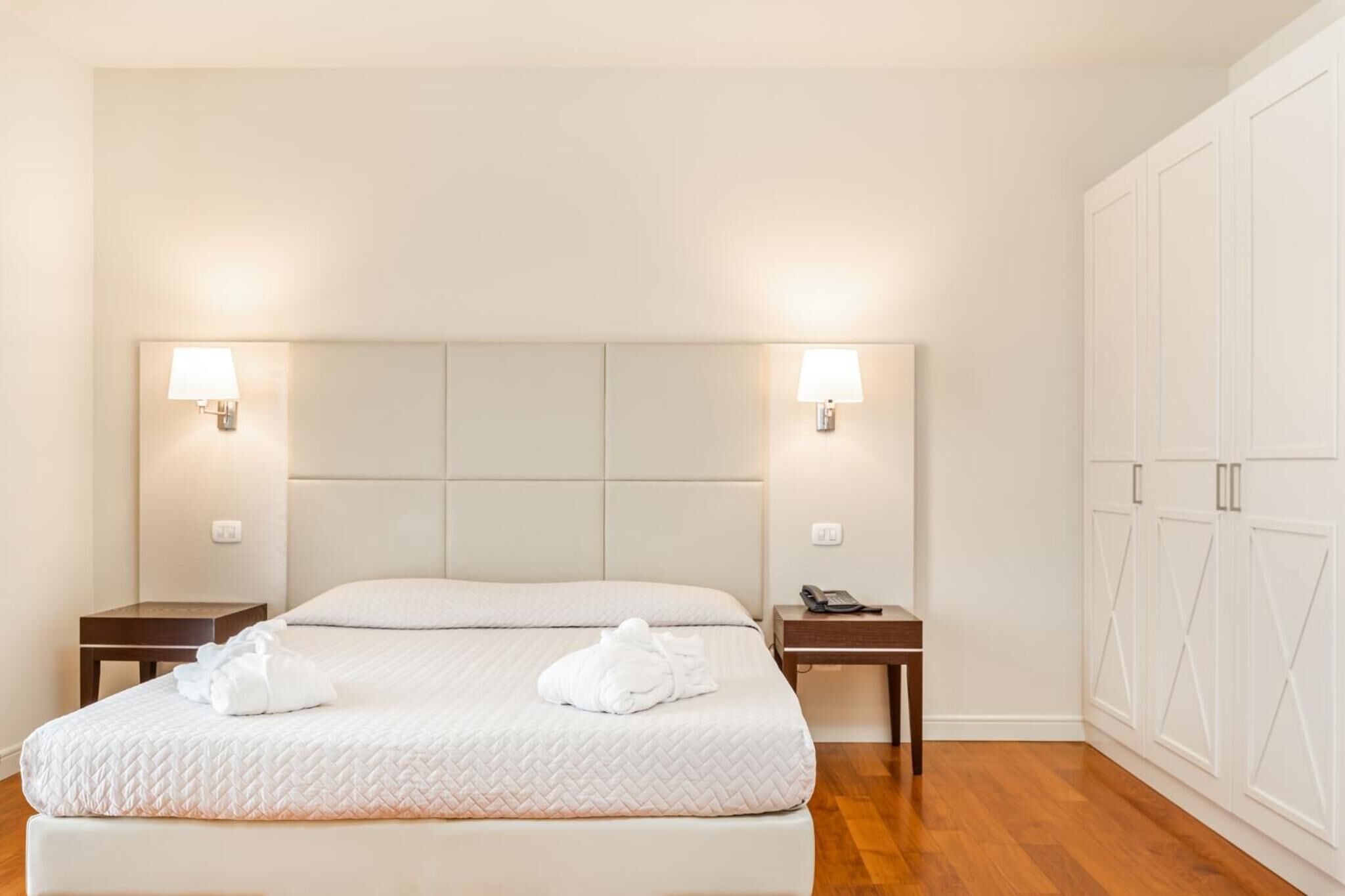 A neatly arranged hotel room with a large bed, two bedside tables, a nightstand, and a wardrobe.