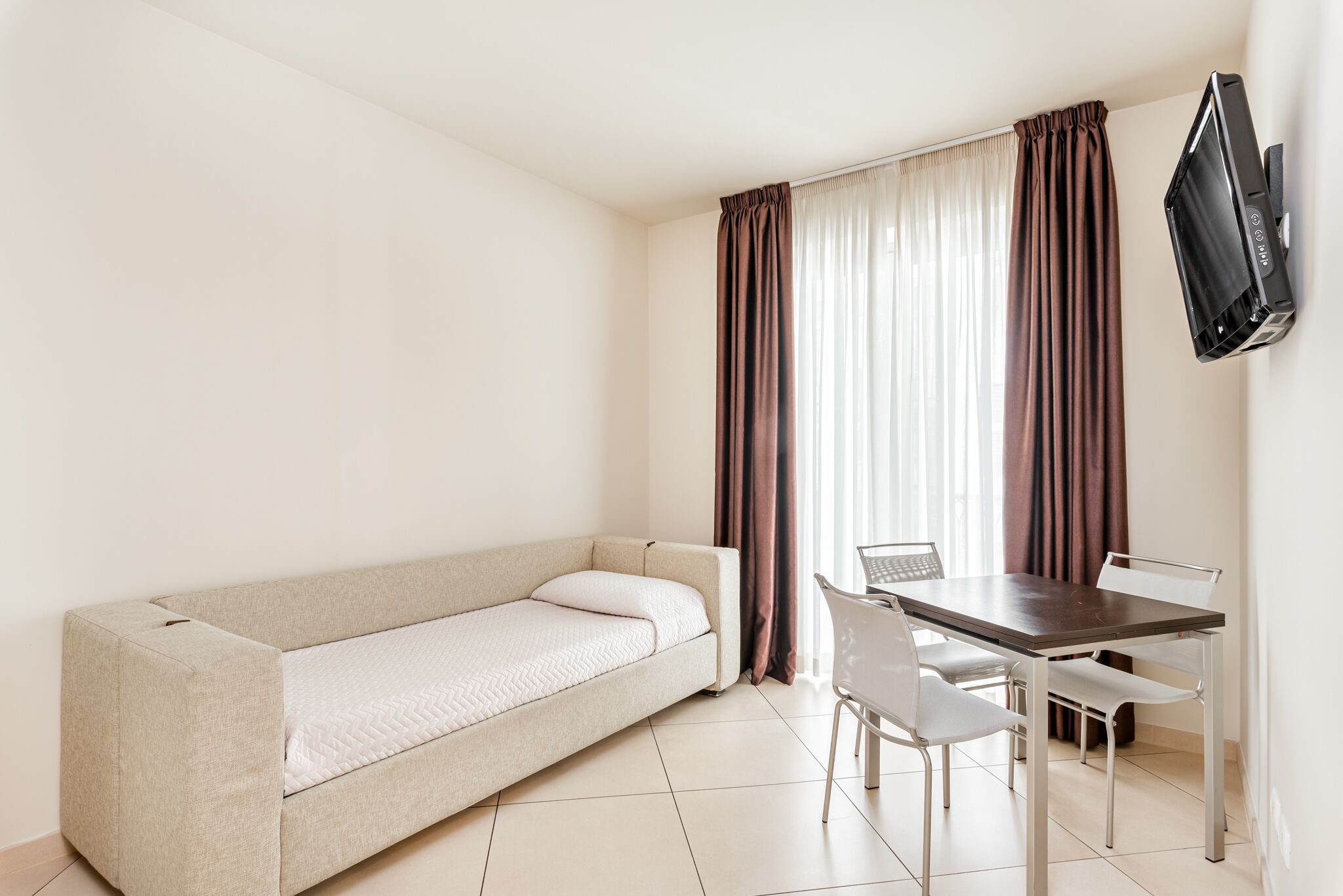 A compact room with a sofa bed, a dining table with chairs, and a TV mounted on the wall.