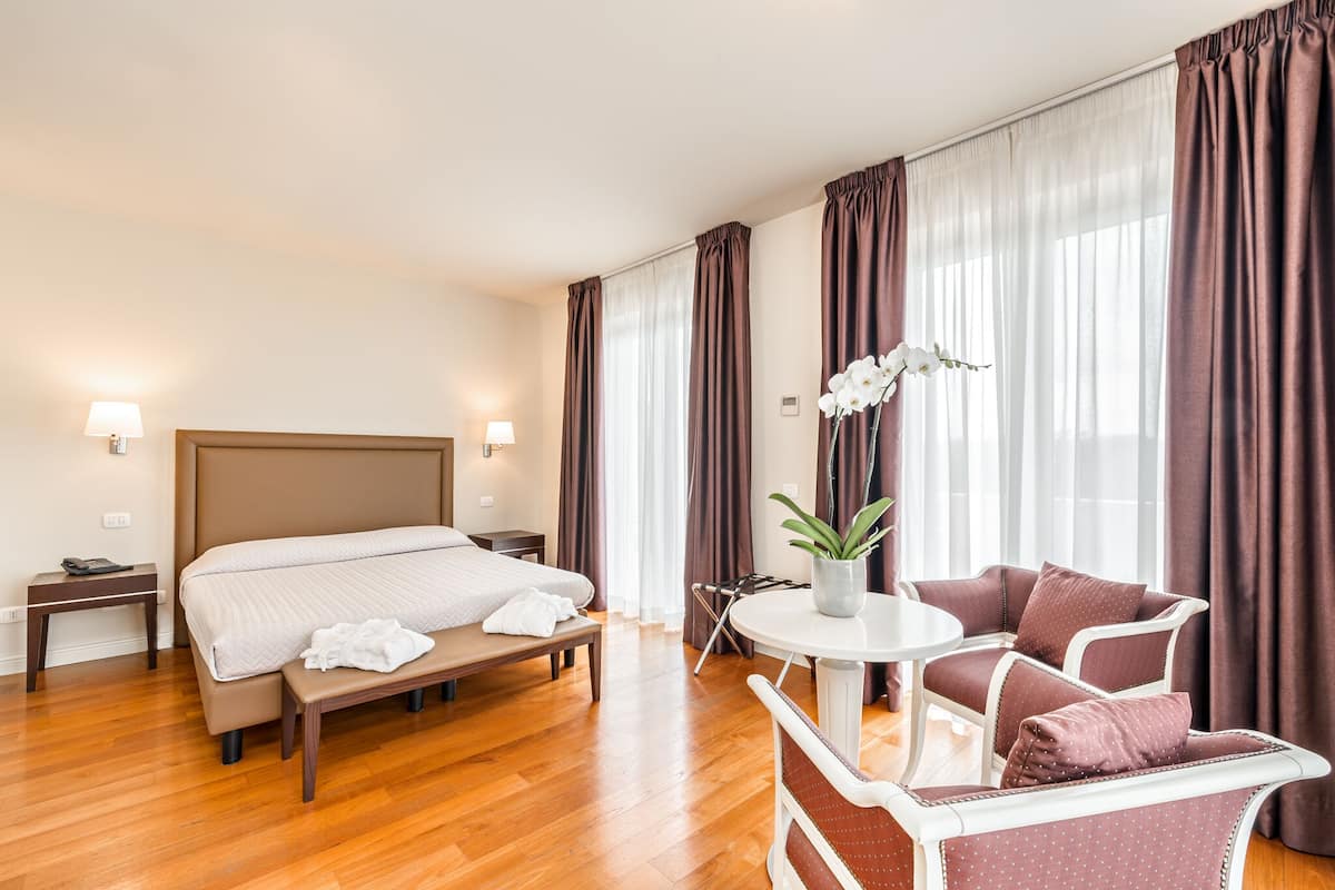 A hotel room with a bed, bedside table, lamp, chair, and a small table with a plant.