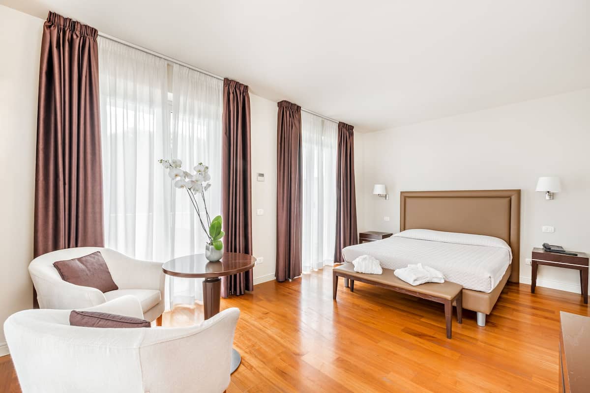 A hotel room with a large bed, a sitting area with a chair and table, and a view through sheer curtains.