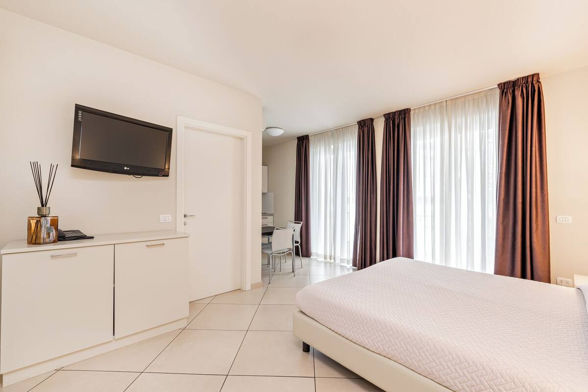 A hotel room with a bed, a TV mounted on the wall, a desk with a chair, and curtains on the windows.